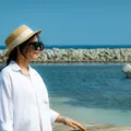 Paduan gaya berpakaian ke pantai dengan kemeja linen putih dan kacamata hitam di bawah terik matahari pesisir.