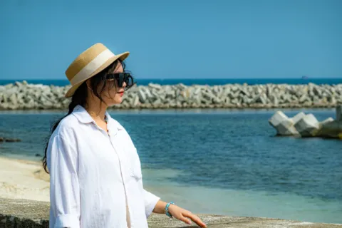 Paduan gaya berpakaian ke pantai dengan kemeja linen putih dan kacamata hitam di bawah terik matahari pesisir.