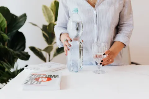 Seseorang sedang menuangkan air minum ke gelas di meja kerja untuk menjaga hidrasi tubuh